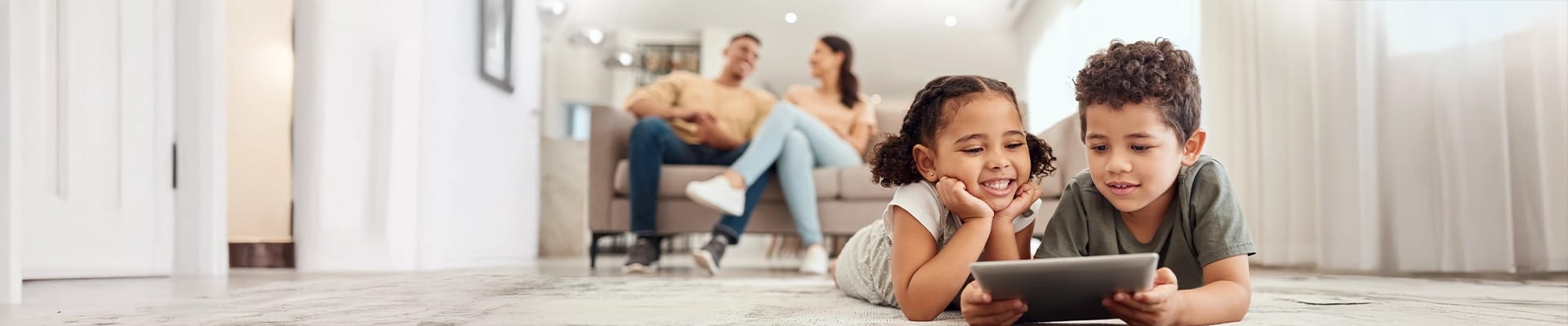 Image of young brother and sister playing on tablet device while father and mother observe in the background